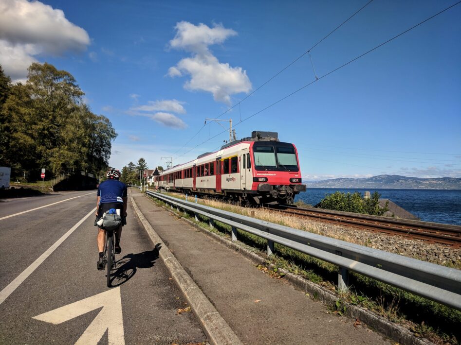 Male cyclist riding along Lac leman with a train travelling in the opposite direction